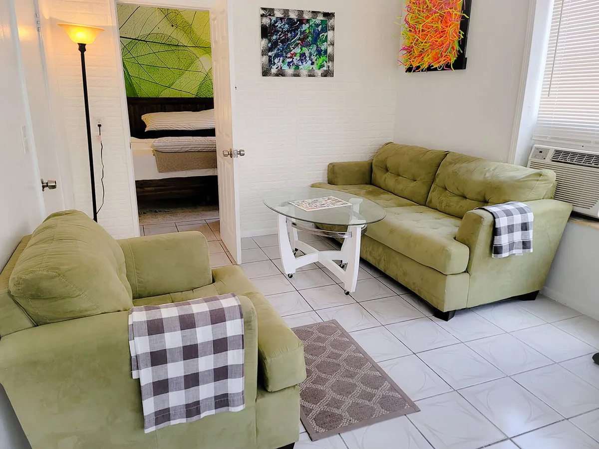 The image showcases a bright, inviting living room with two olive green sofas draped in plaid throws, a sleek glass coffee table, and colorful abstract artwork that energizes the space. An open door reveals a bedroom adorned with a large, lush leaf mural, hinting at a tranquil retreat beyond the main living area.
