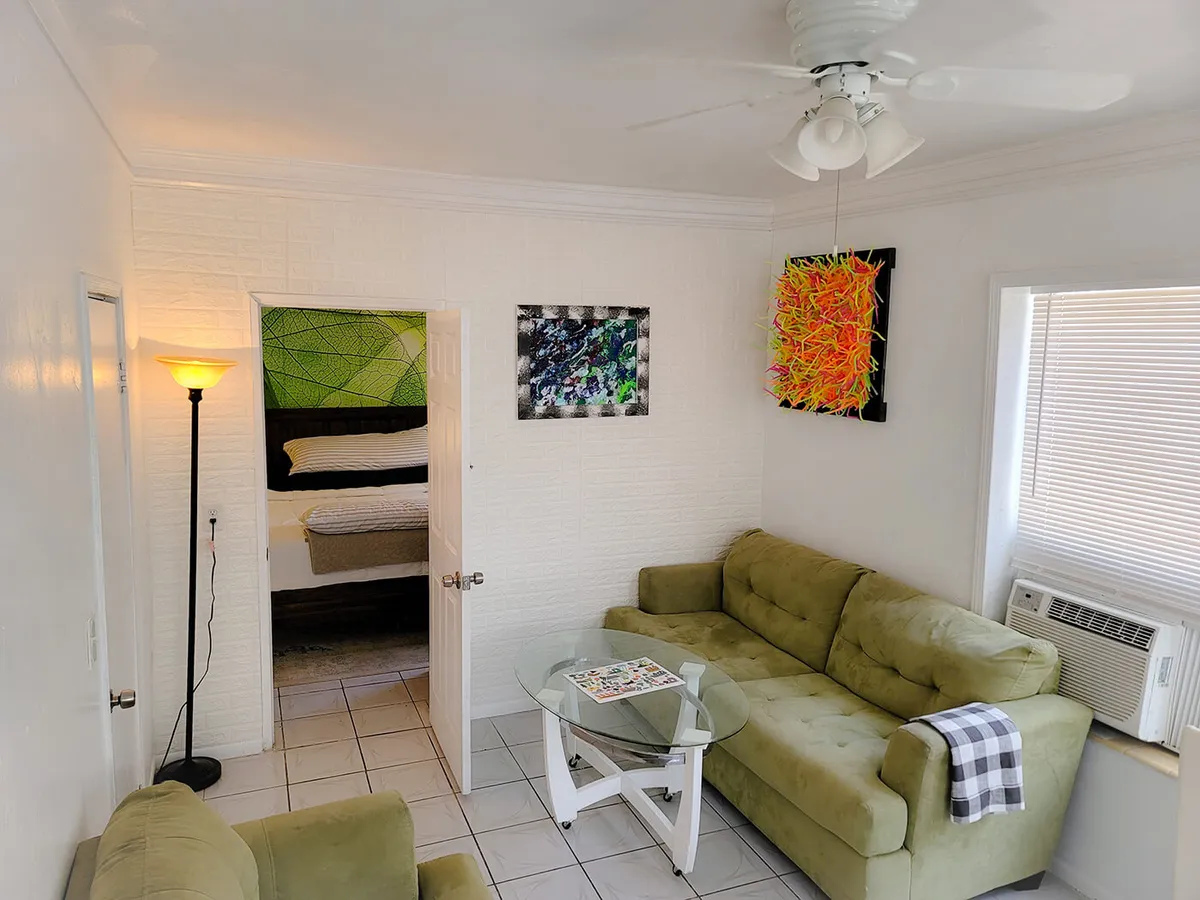 The cozy living room features a green tufted sofa with a plaid throw, a glass coffee table with a magazine, and colorful abstract wall art. There’s a glimpse into the adjacent bedroom with a green leaf mural, tying together the open-concept flow and connecting living and resting spaces.