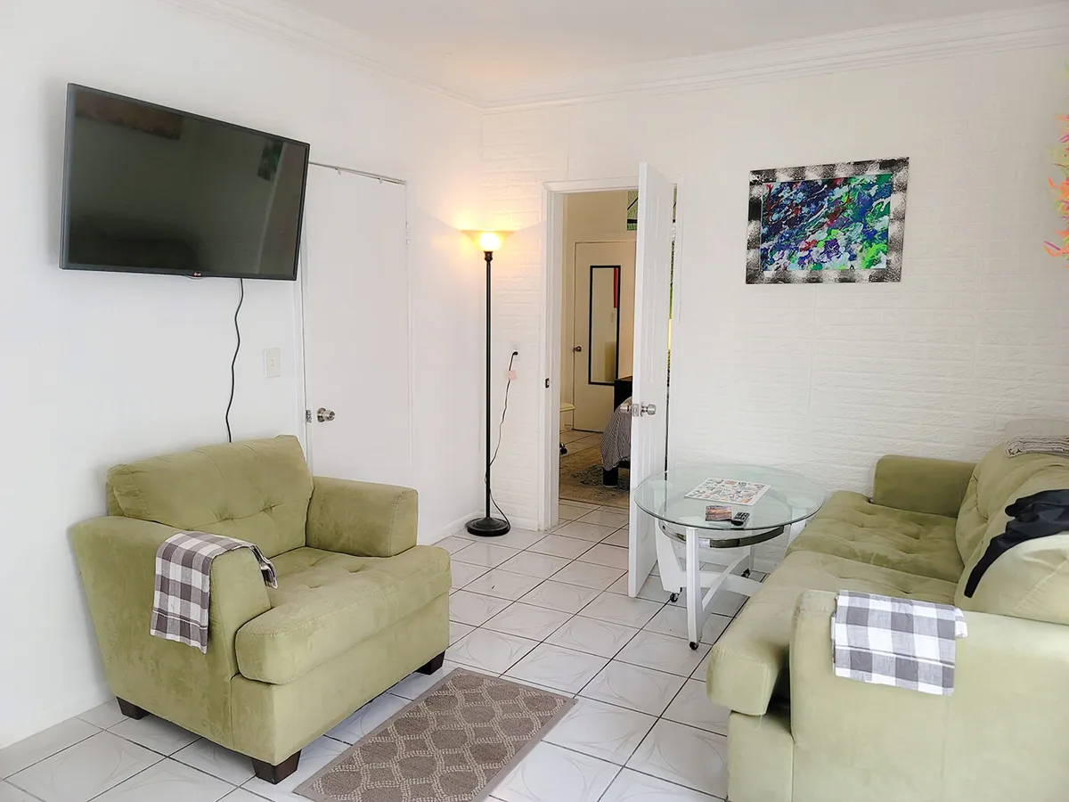 A green armchair with a plaid throw sits beside a glass coffee table and a wall-mounted TV, with a floor lamp illuminating the space. The room is bright, airy, and welcoming, ideal for relaxing or catching up on work with high-speed WiFi.