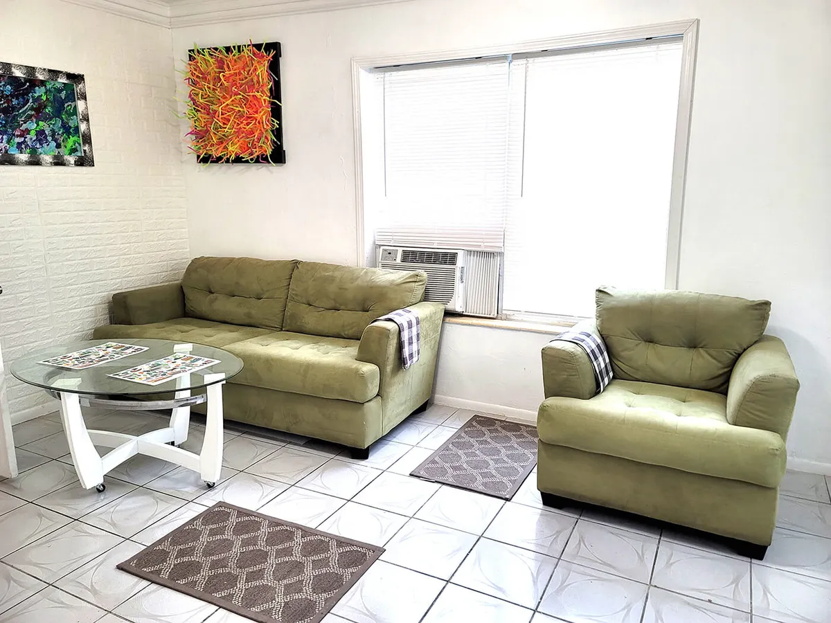 A bright and comfortable living area centered around a green upholstered sofa and a matching armchair. A glass coffee table with a white base sits at the center, with colorful abstract wall art adding personality to the space. The arrangement suggests a welcoming spot for socializing, lounging, and prepping for outings or unwinding after events.