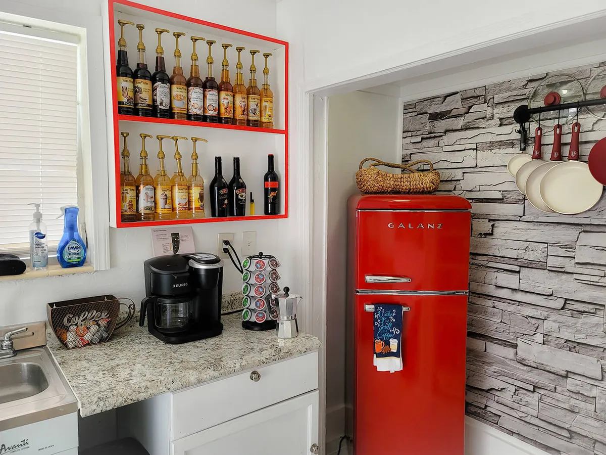 The photo spotlights a bright red vintage-style refrigerator, a dedicated coffee station with a Keurig coffee maker and pods, wall-mounted syrup dispensers, and a pan and utensil rack set against textured stone-patterned walls. The setup is lively and practical, perfect for brewing fresh coffee and preparing simple meals.