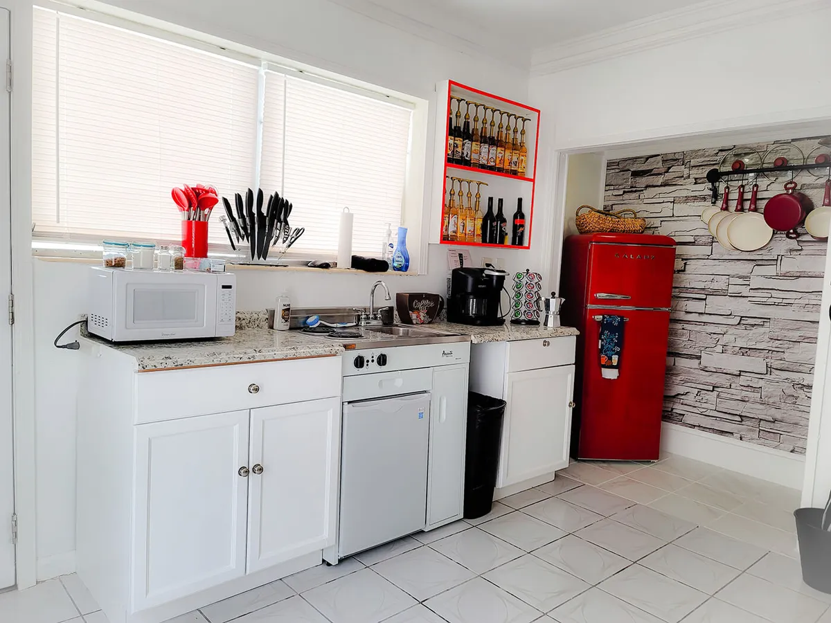 This image features a bright red retro-style refrigerator as the focal point, complemented by a white granite countertop with various kitchen appliances. Open shelving displays bottles, and hanging pots and pans add functionality and character against a stone-patterned wallpaper backdrop. The scene embodies a practical, inviting space ideal for meal preparation and coffee breaks.