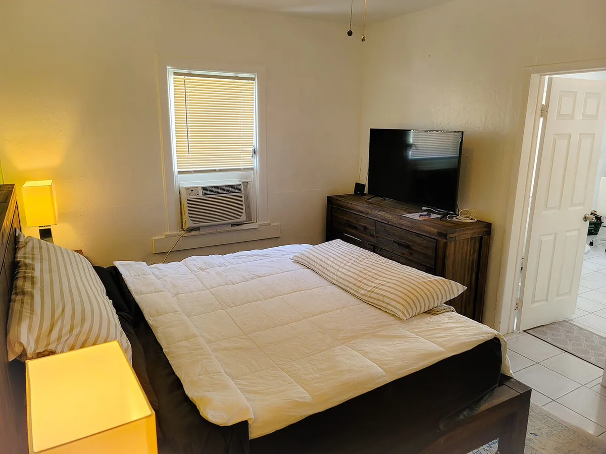 A cozy bedroom featuring a king size bed with a white comforter and striped pillows, flanked by a wooden dresser with a flat screen TV. The room is well-lit by a window with blinds and equipped with an air conditioning unit and a bedside lamp, contributing to a comfortable and inviting atmosphere.