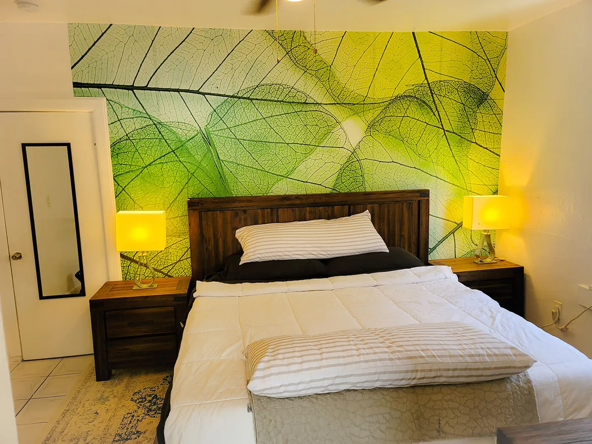 A bright, inviting bedroom showcasing a king-sized bed with white comforter and striped pillows, set against a bold green leaf mural. Two wooden nightstands with matching yellow lamps add symmetry, while a full-length mirror on the door offers practical convenience.
