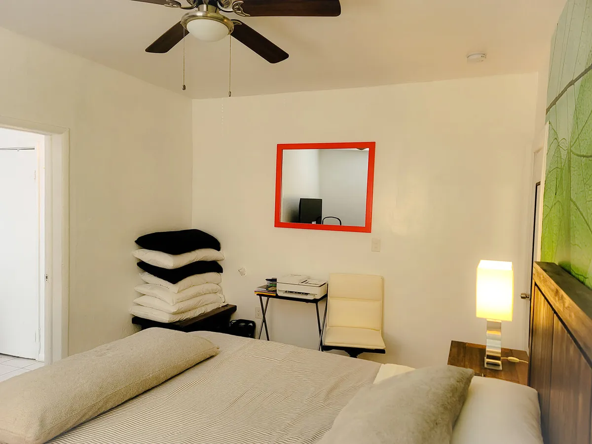 A cozy bedroom scene with a comfortable king bed adorned in neutral bedding, complemented by a stack of extra pillows and blankets. The room is thoughtfully arranged with a red-framed mirror on the wall and a compact desk setup with a chair and lamp, suggesting both rest and productivity are possible in this adaptable space.