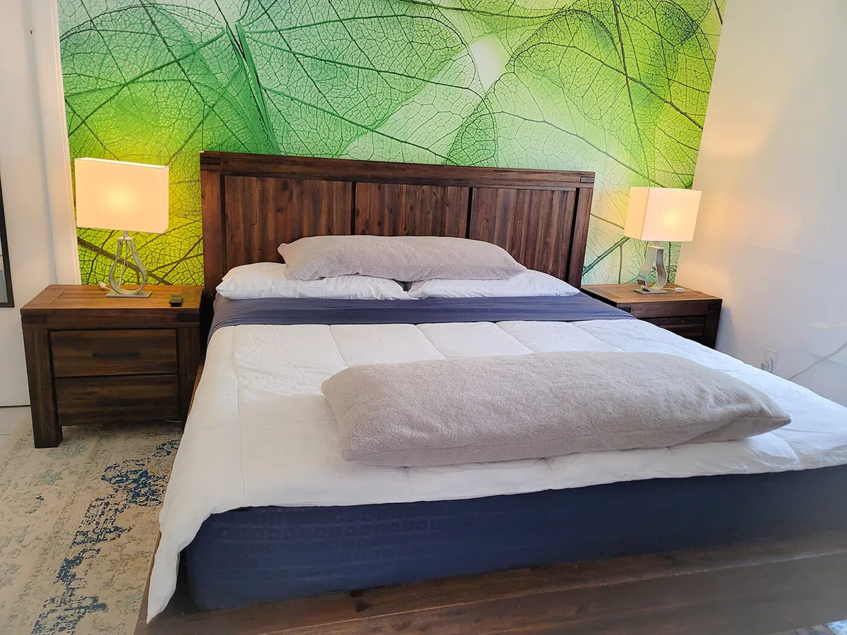 A serene bedroom setting featuring a king-sized wooden bed dressed in crisp white and blue linens, centered against a lively leaf-patterned green accent wall. Matching wooden nightstands with soft-glow lamps flank the bed, while a plush rug peeks out beneath, contributing to a tranquil and inviting atmosphere ideal for relaxation and restful sleep.