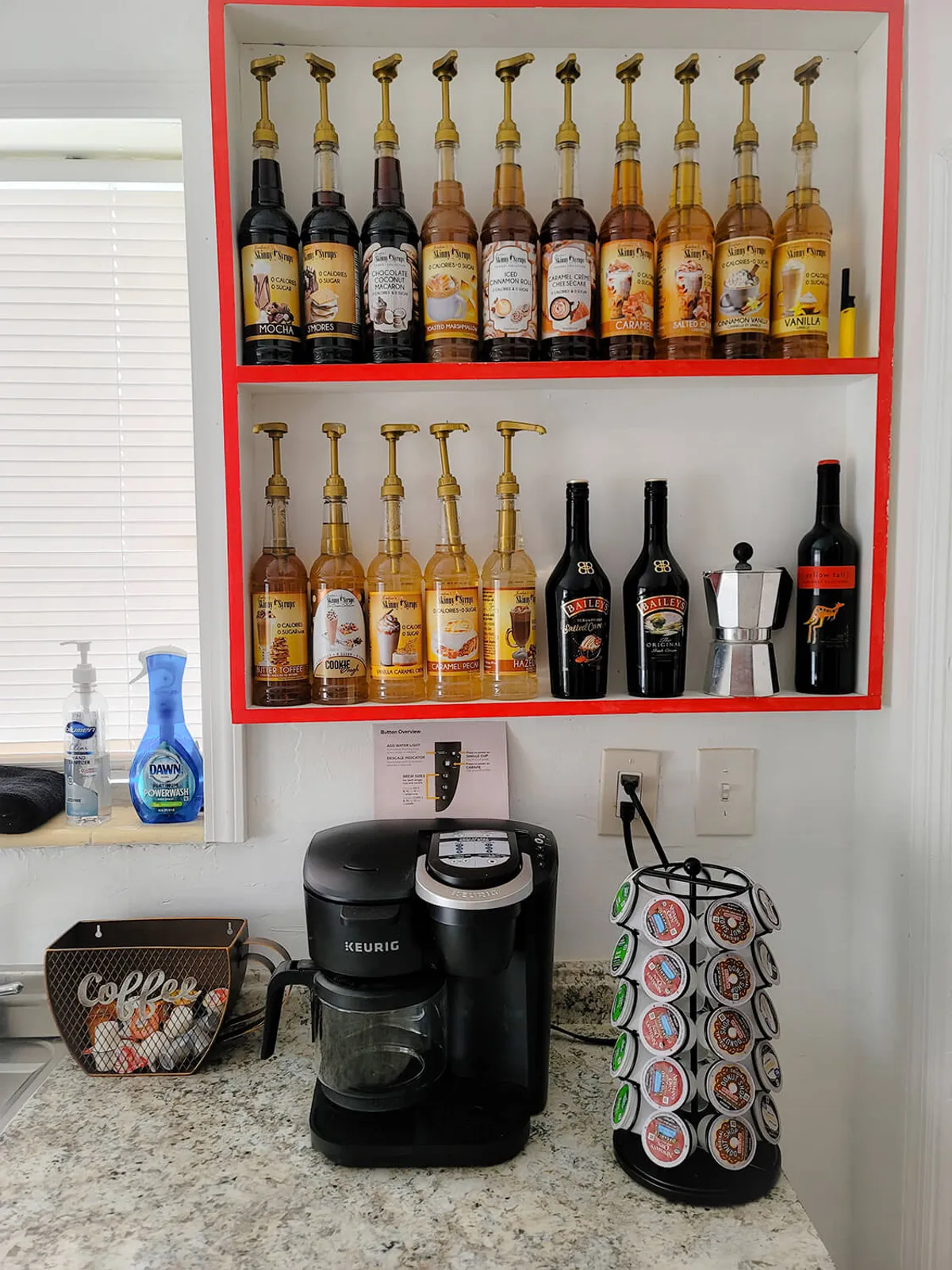 Red shelves display a variety of coffee syrups above a Keurig coffee maker, a rotating coffee pod holder, a small moka pot, and bottles of Baileys. The scene is cozy and encourages guests to customize their morning coffee routines.