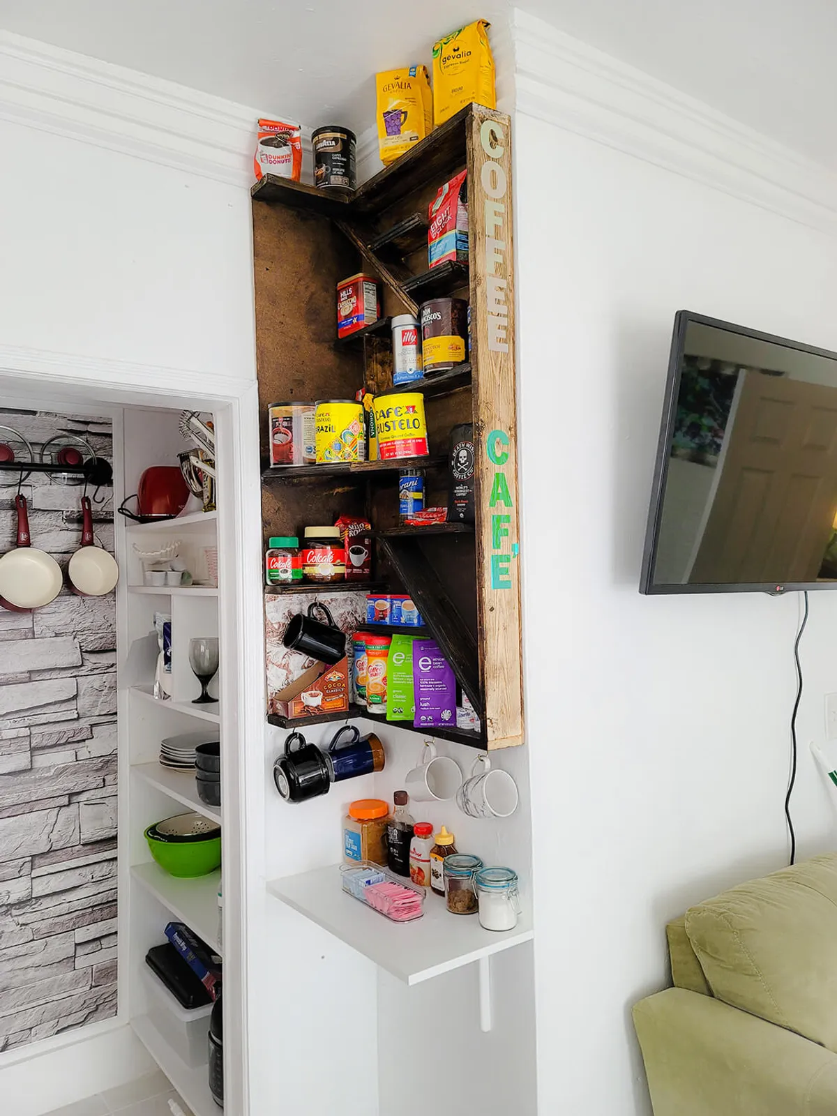 A coffee shelf stocked with a variety of coffee brands and mugs sits above a small white counter with supplies, next to an open pantry with kitchenware. A wall-mounted television is visible, suggesting the convenience of enjoying a drink while relaxing or socializing nearby.